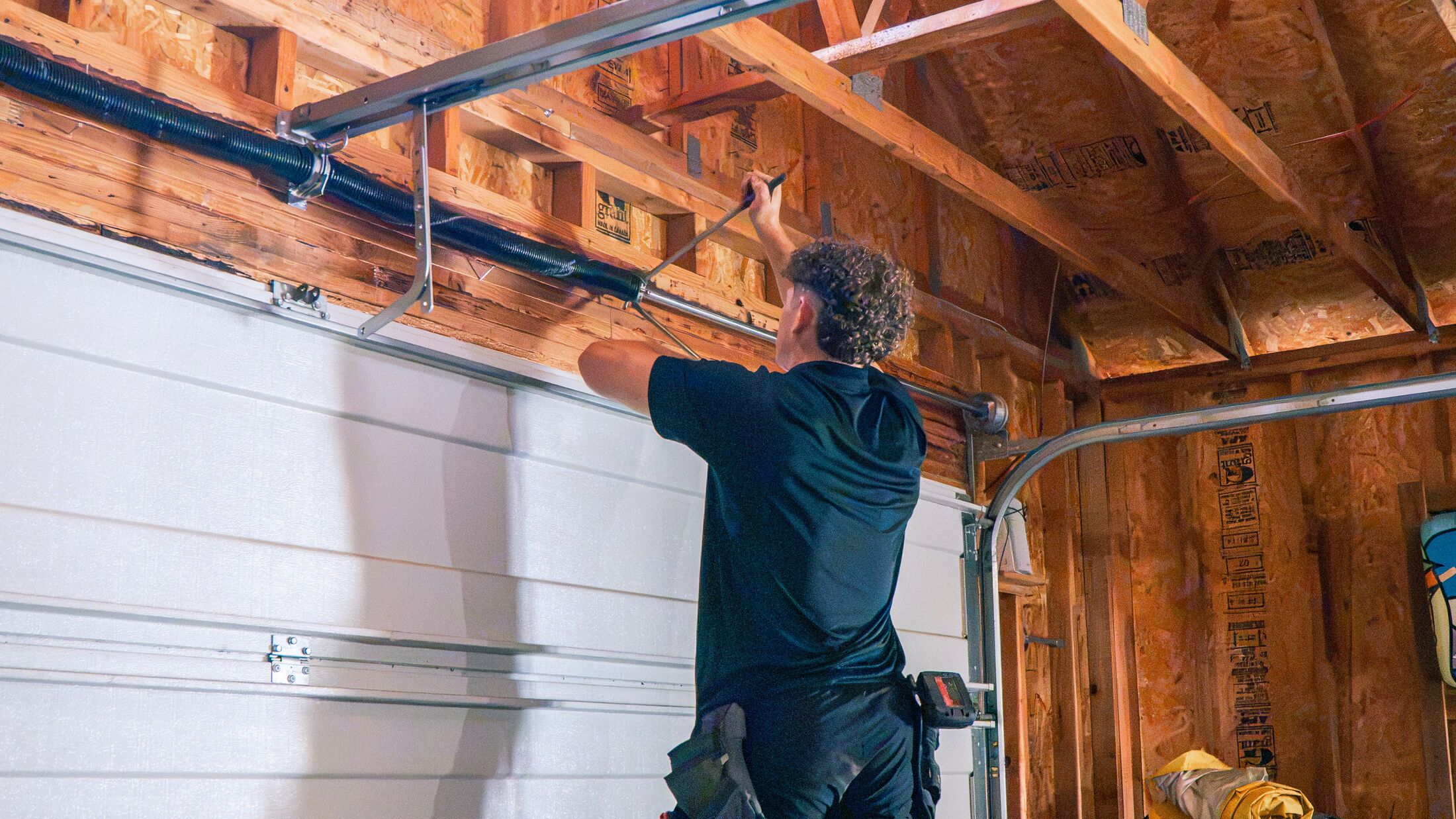 An Elevation technician on a ladder working along the track of a garage door, tools laid out below